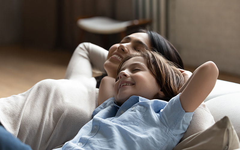 A woman and a child are resting comfortably together on a couch, smiling, with their hands behind their heads.
