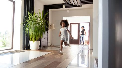 A smiling young girl runs through her family’s living room while her mother watches from a doorway at the back of the room.